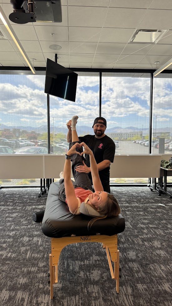 Shawn Taylor stretching a client on a table in an office while she makes a heart with her hands