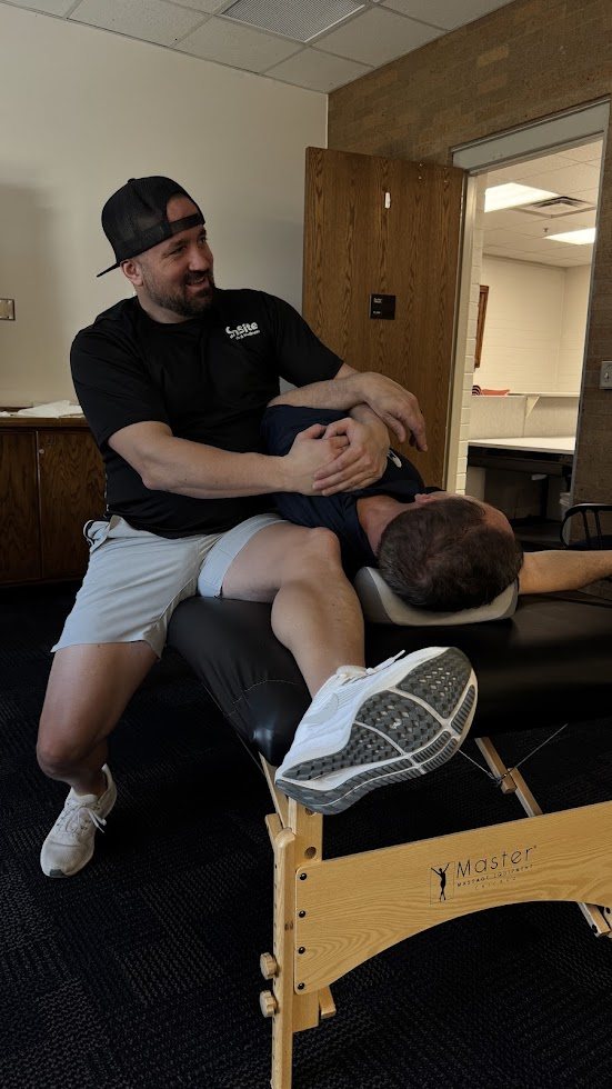 Shawn Taylor sitting on a stretch table, smiling while stretching a male client's hip